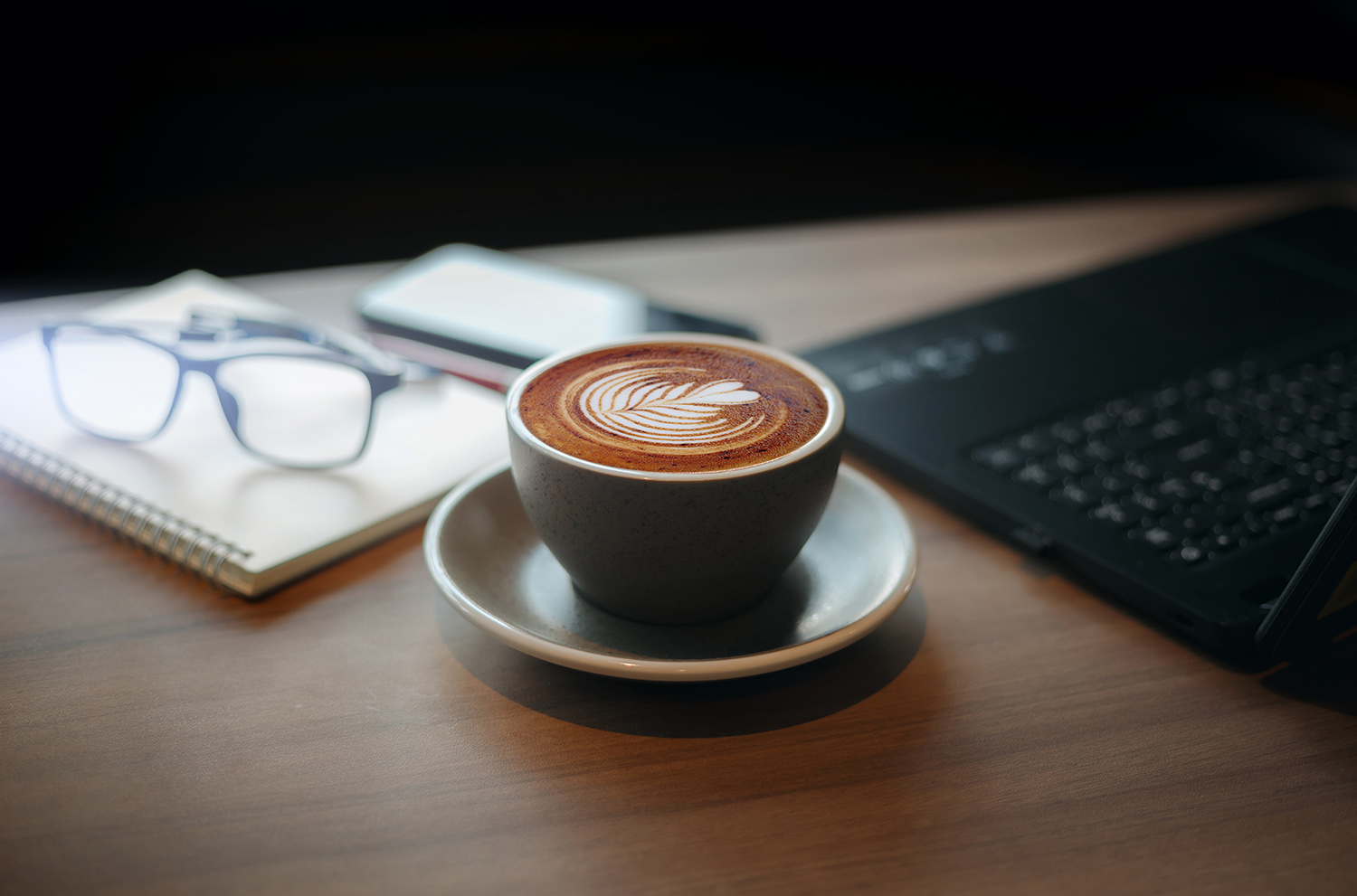 Close-up view, white cup of coffee with computer laptop, notebook, pen and eye glasses on wooden table in cafe, People resting after work in a calm cafe